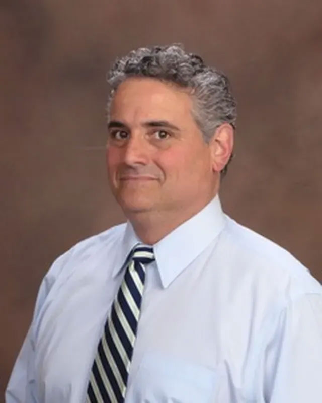 The image depicts a man with a bald head, wearing a blue tie, standing against a beige background.