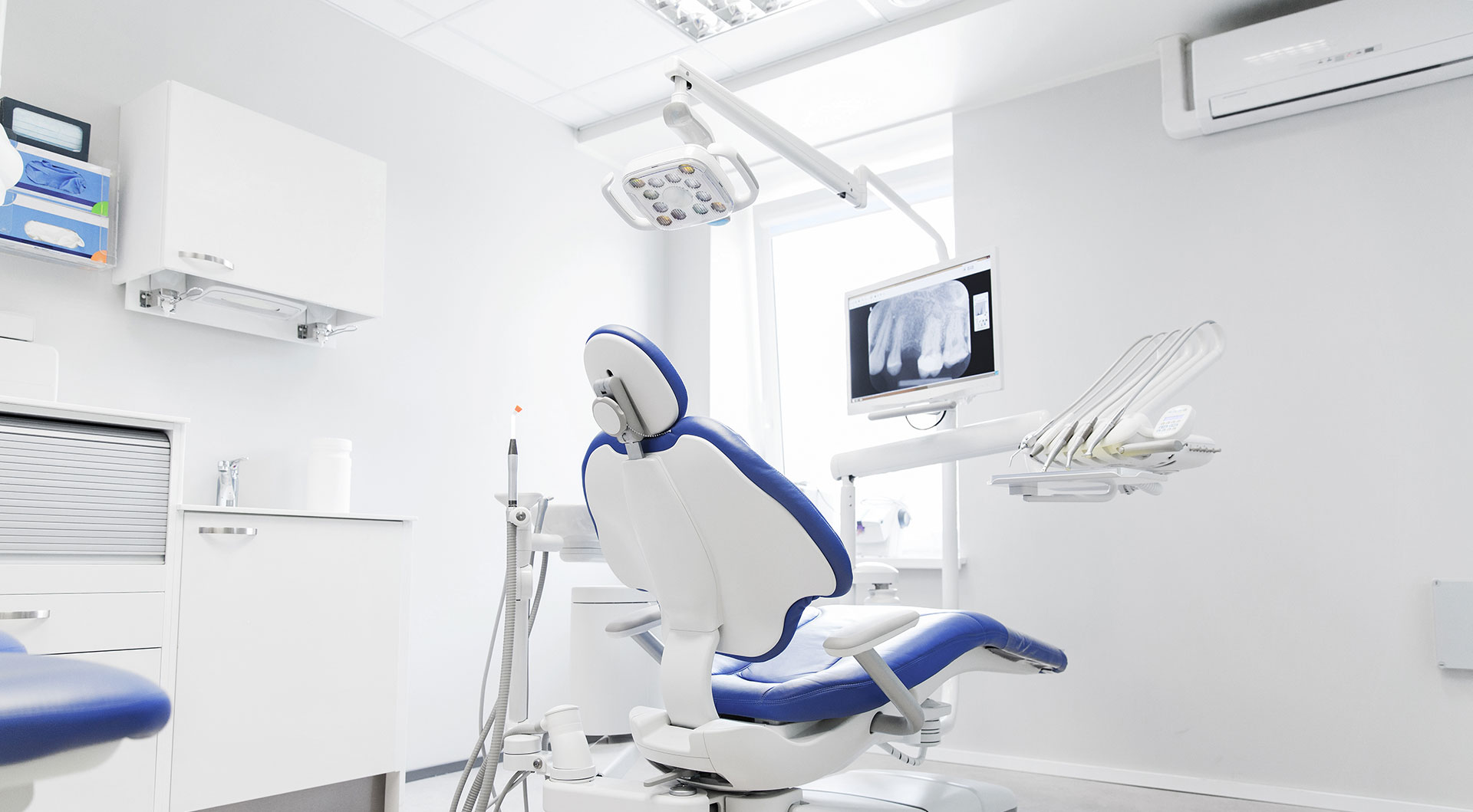 The image shows an interior view of a dental practice with a modern chair and equipment, including a computer screen displaying a patient s X-ray, set against a clean, white background.