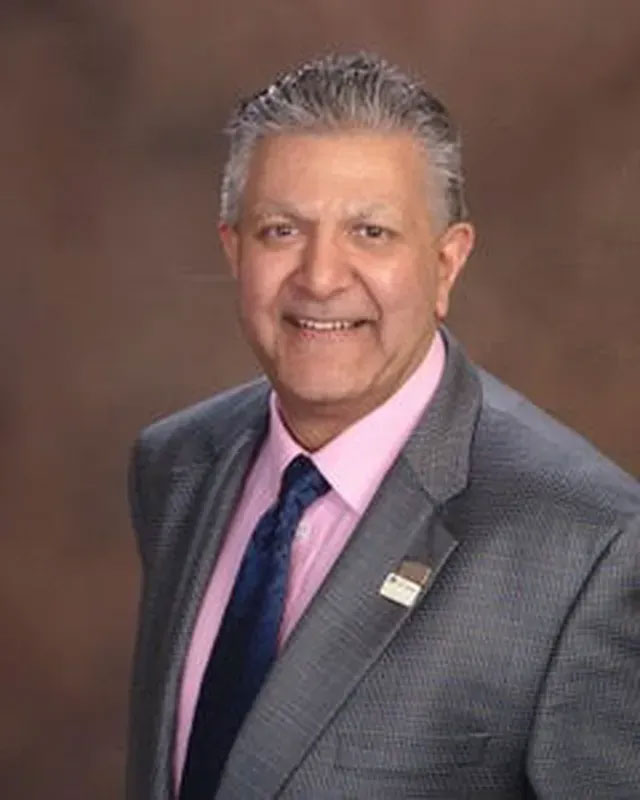The image shows a man standing against a background with a name badge, wearing a suit and tie, smiling at the camera.