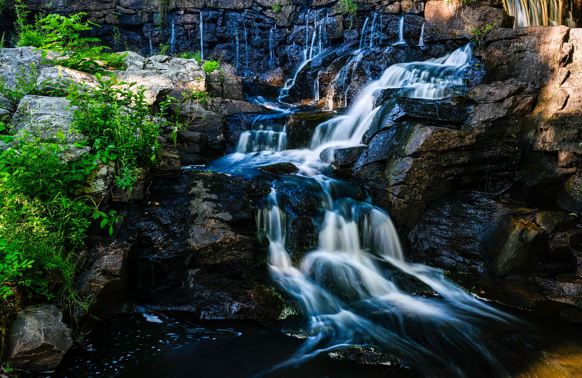 The image shows a serene natural scene with a waterfall flowing into a pool, surrounded by lush greenery and rocks, set against a clear sky.